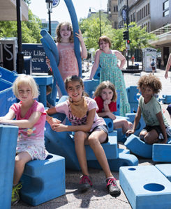 A group of six children sitting and standing near a pile of large foam blocks outdoors on a sunny day 