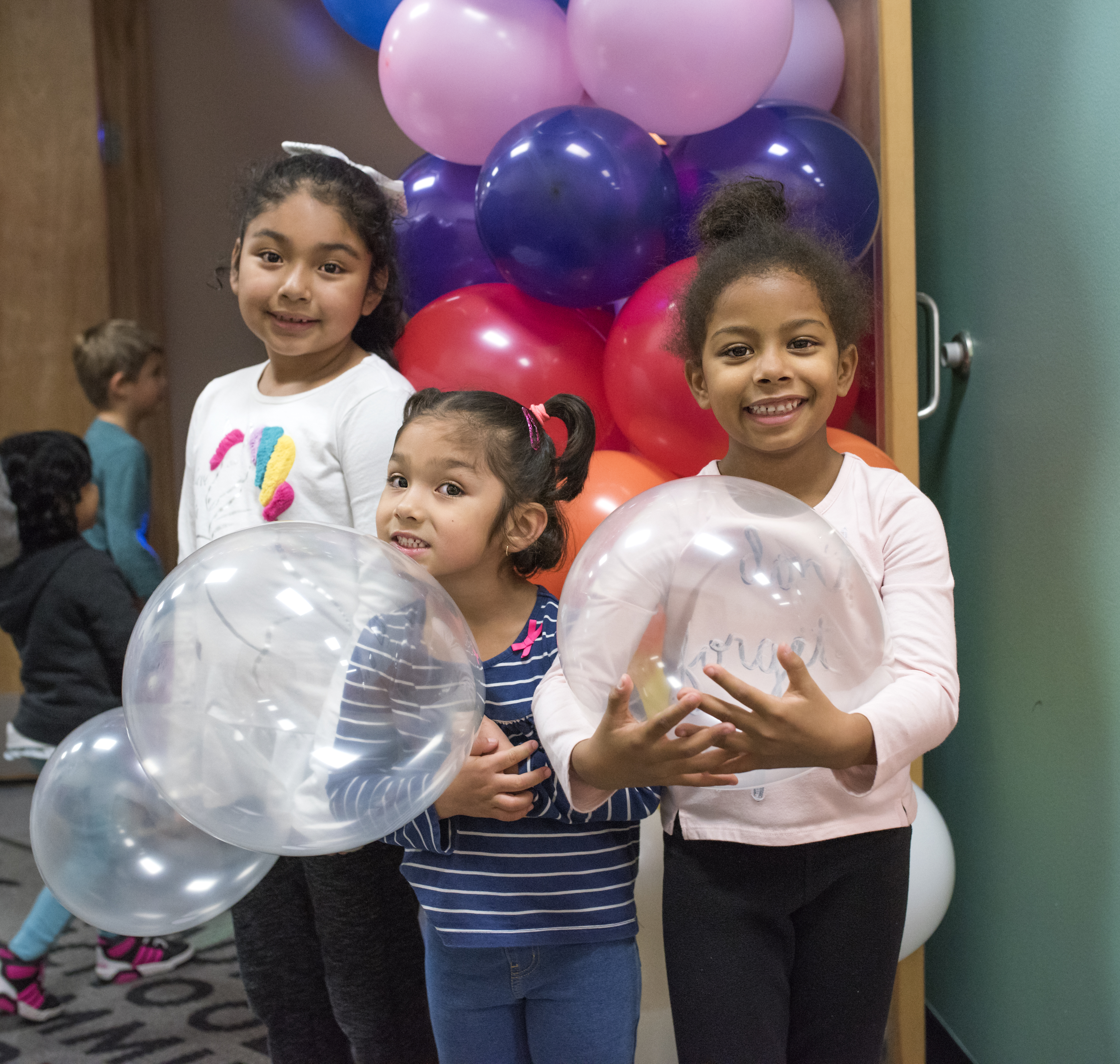 Three little girls of color holding balloons and standing in front of a column of balloons