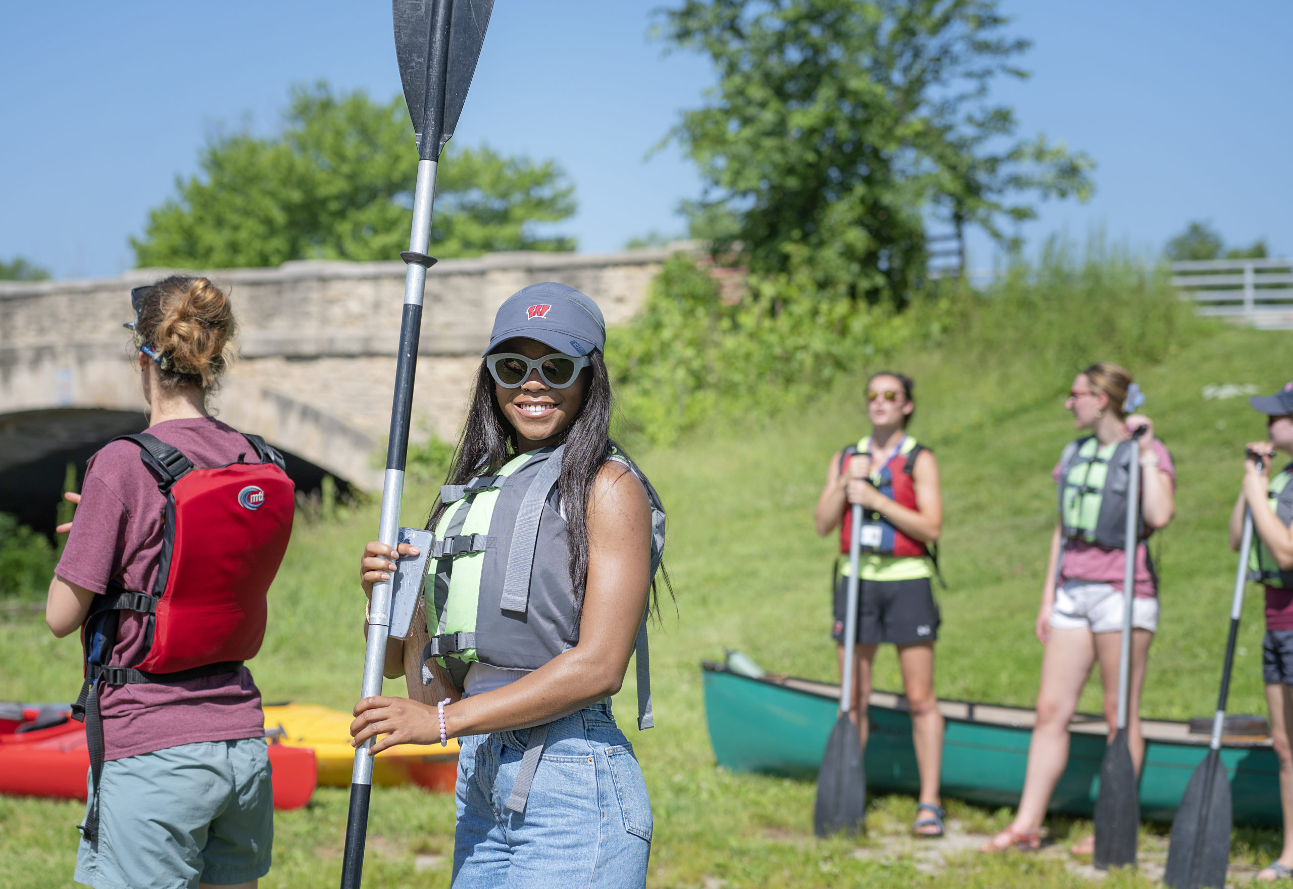 A group of people getting ready to go canoeing together on the bank of a river near a stone bridge on a sunny summer day