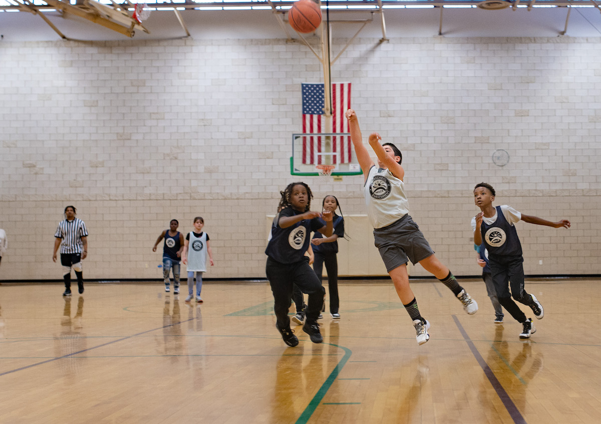 A group of middle schoolers play basketball on an indoor court. One player is airborn as he shoots a basket.