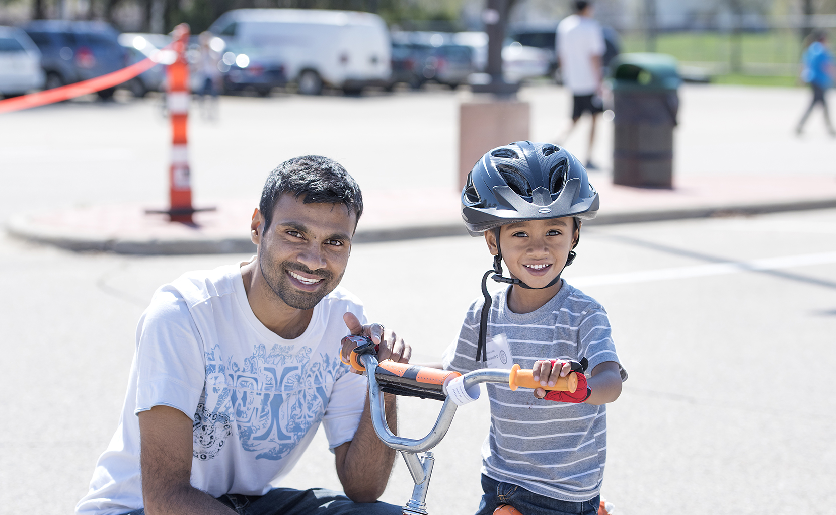 A man is crouching down next to a small child that is sitting on a bike with a helmet on the blacktop or a parking lot