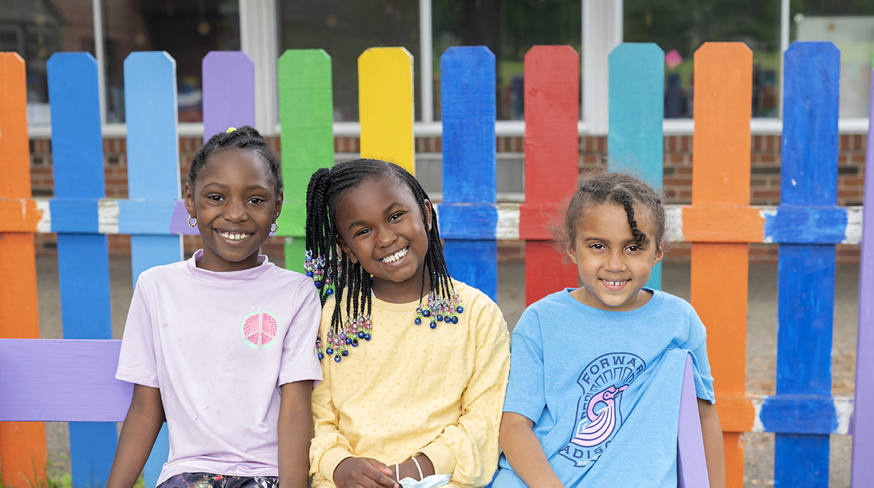 Three little girls of color sitting on a bench in front of a rainbow painted picket fence