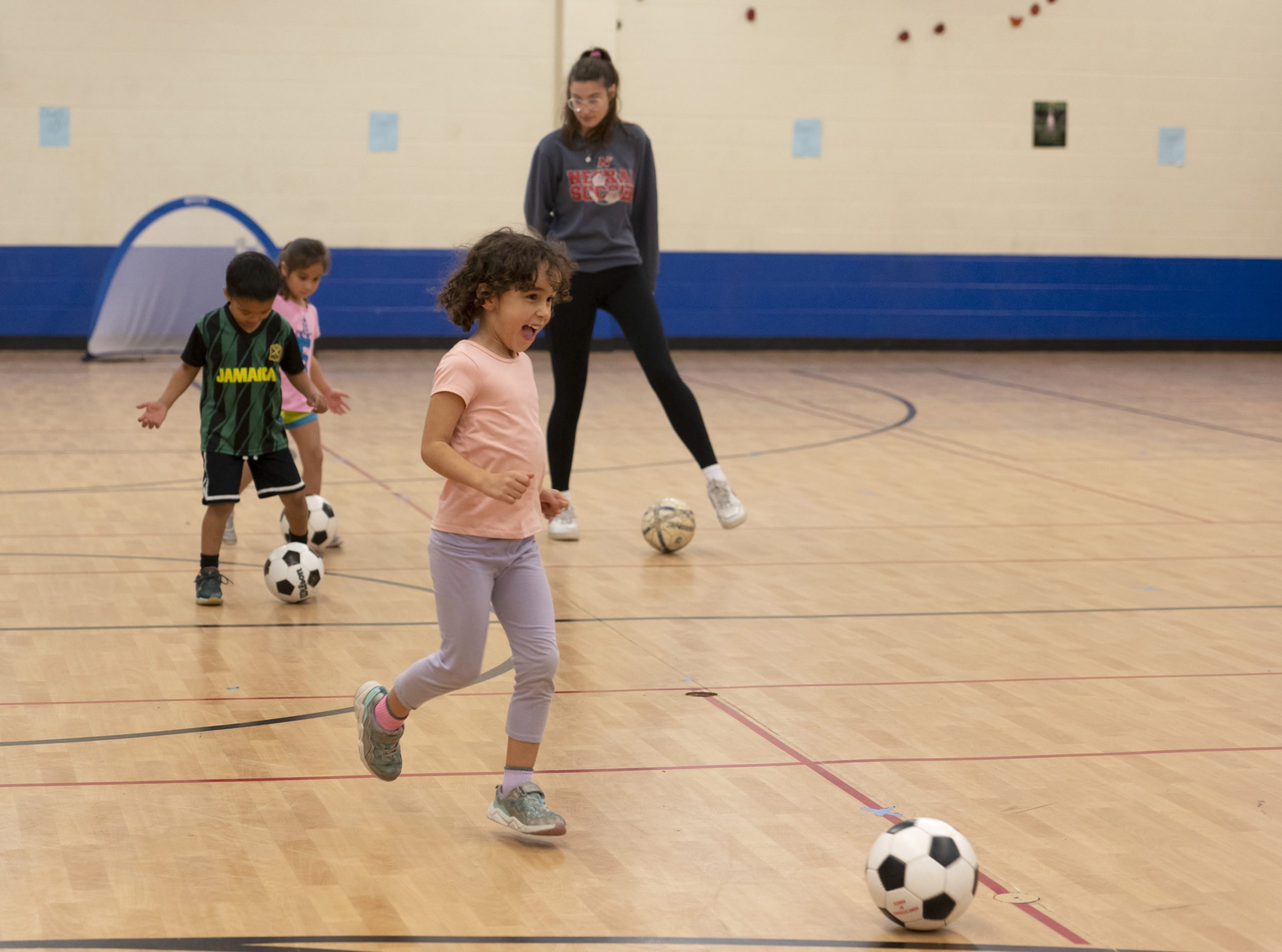 Small kids practicing soccer in a gymnasium while a coach looks on