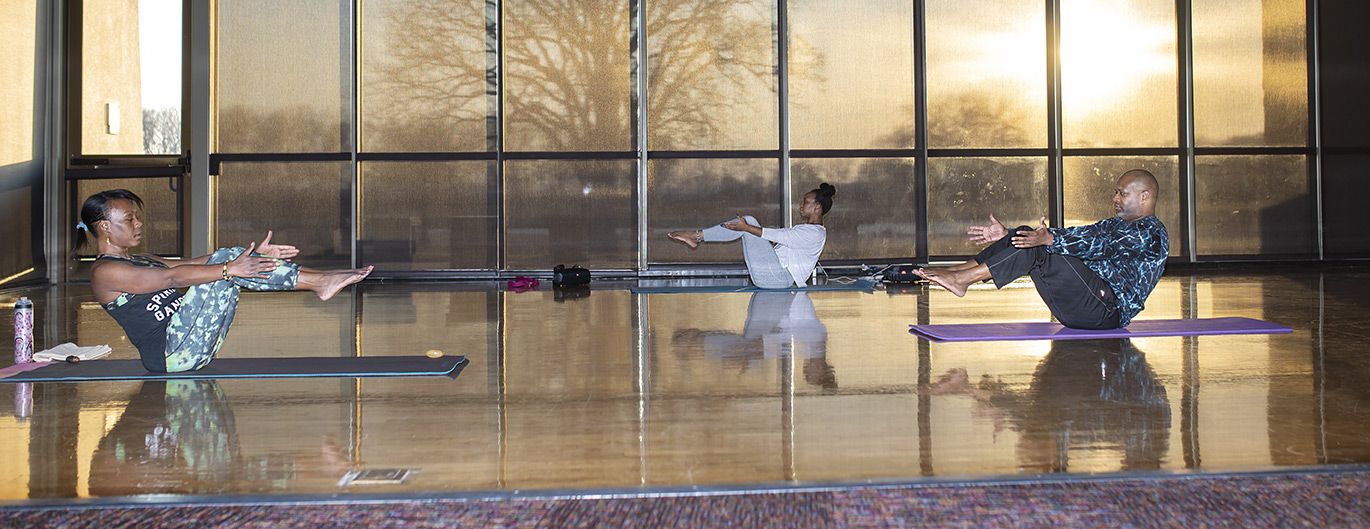 Three people of color doing yoga in a large room with floor to ceiling windows as the sun rises