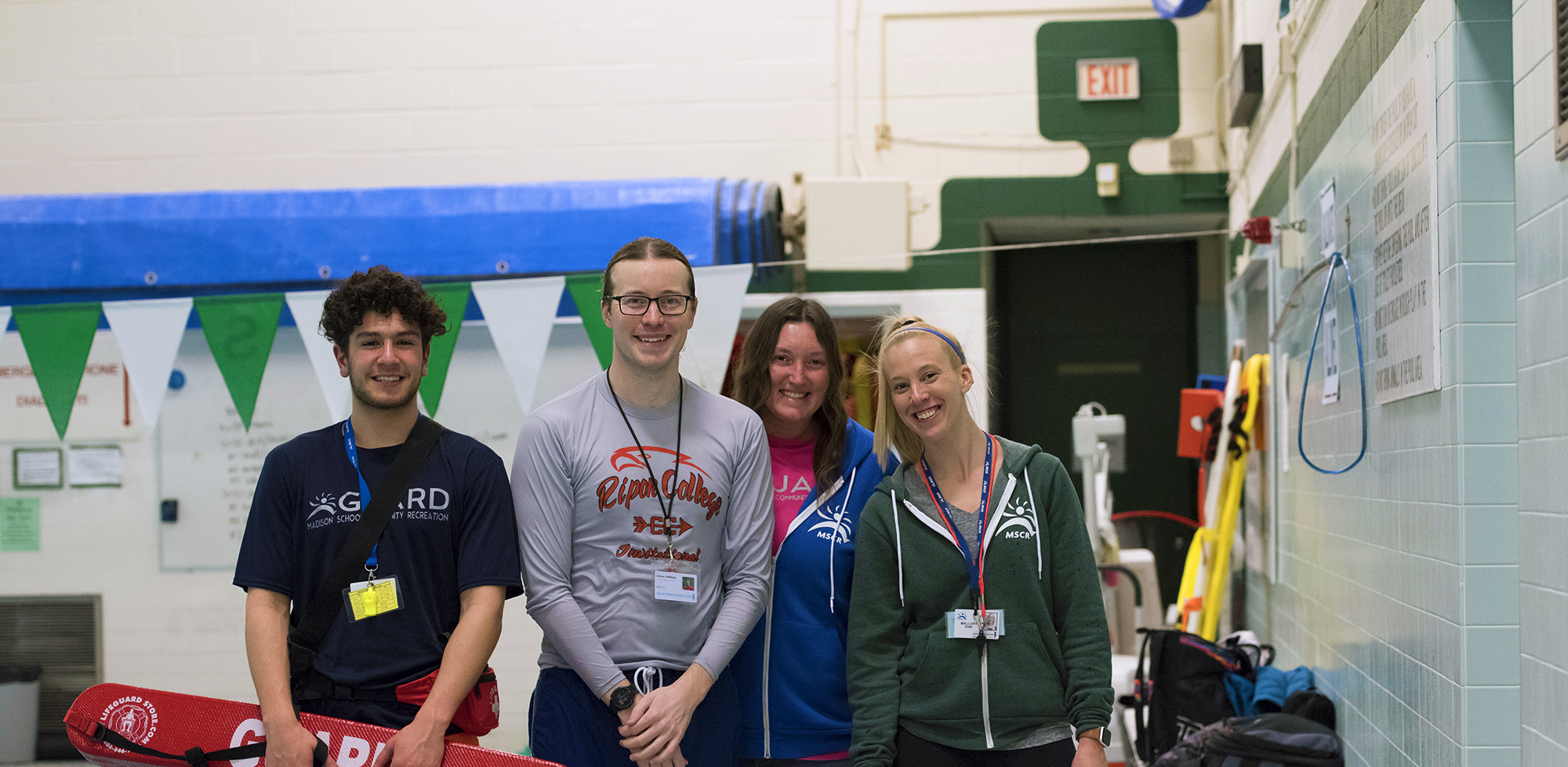 Four young adults standing in a row next to a public indoor pool