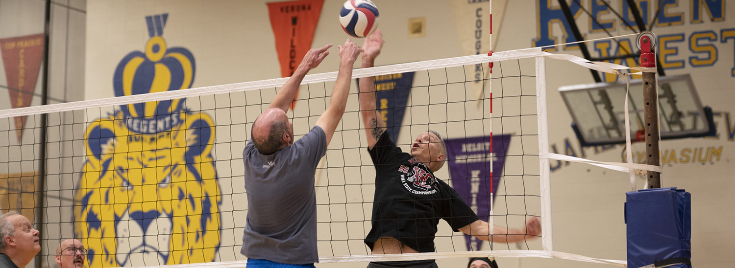 Two men on opposite sides of the volleyball net are vying for the ball, jumping in the air, in a gymnasium